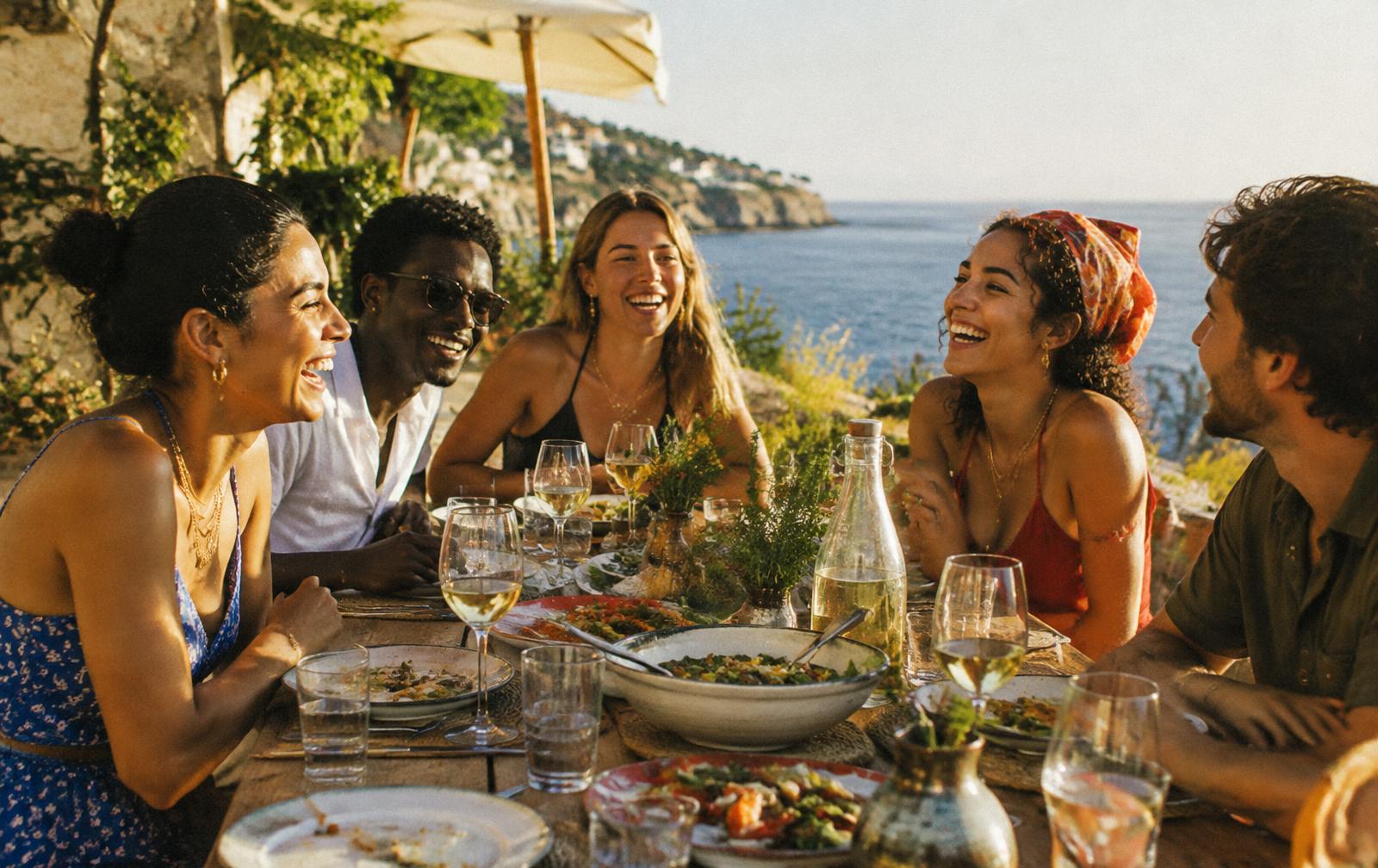 Friends laughing over a long dinner on a sunny seaside terrace