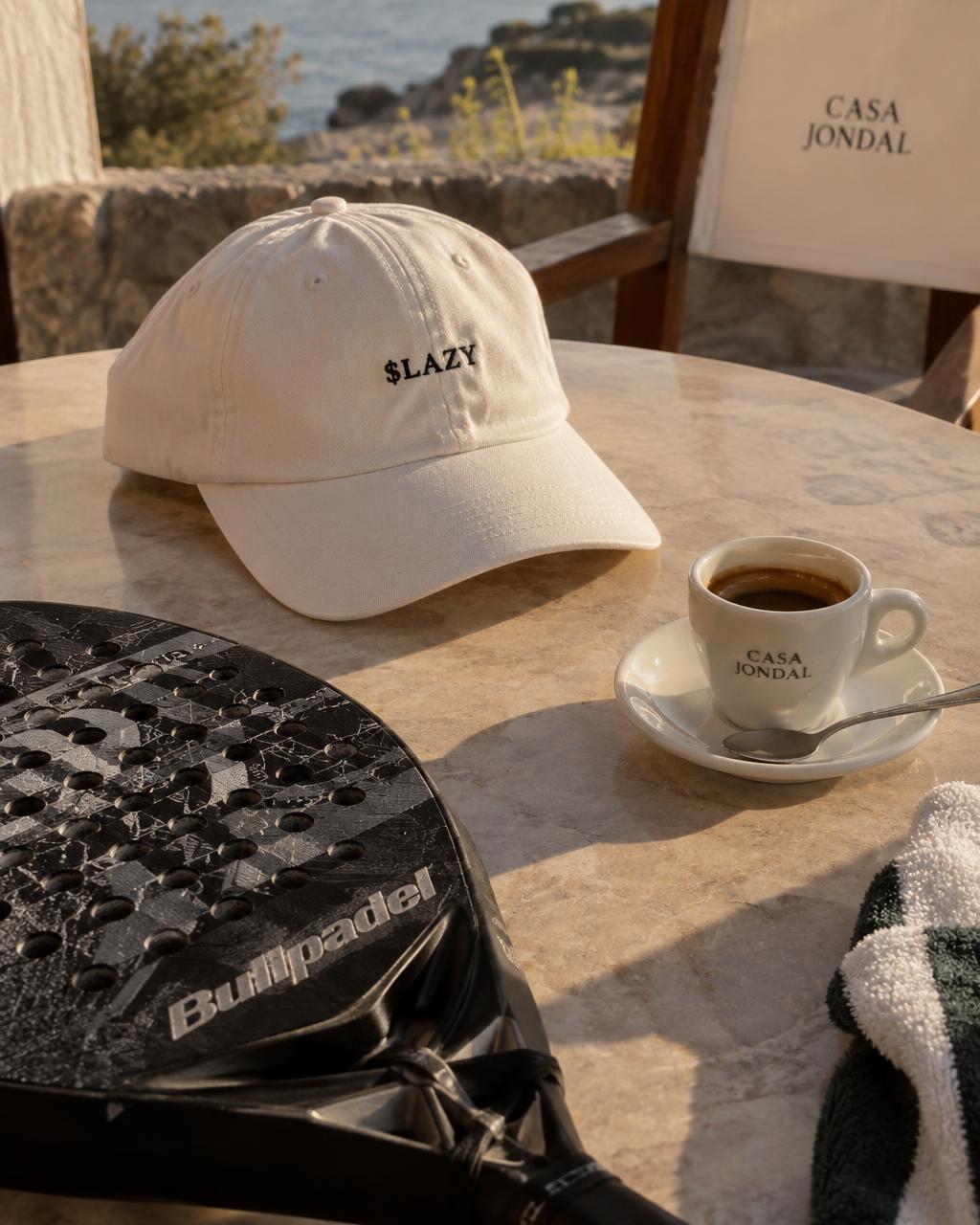 A cream $LAZY cap on a sunny café table beside an espresso