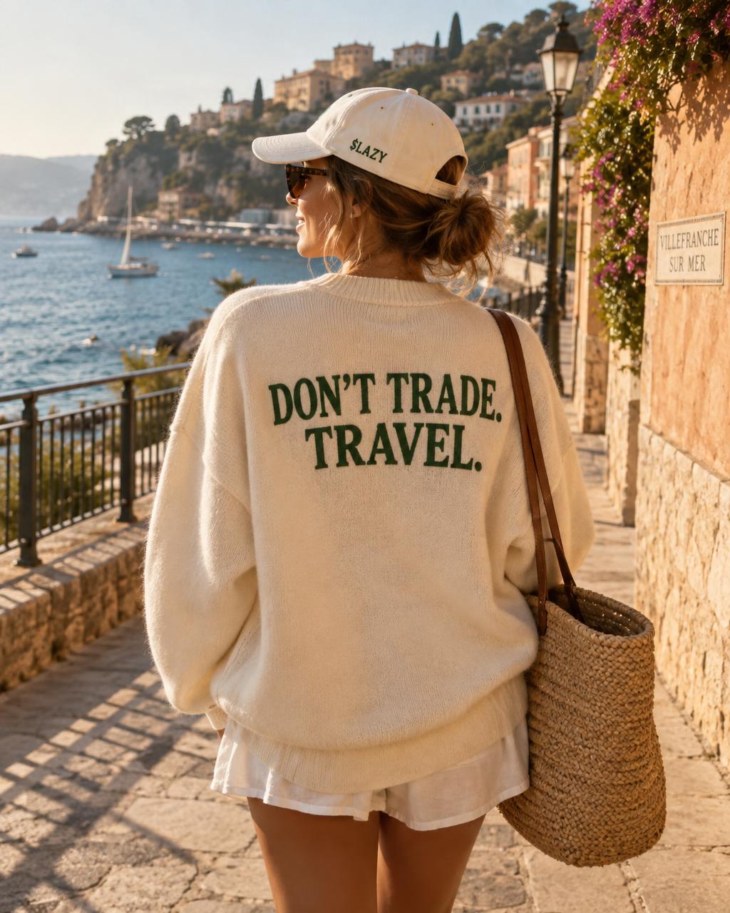 A woman in a cream $LAZY cap and a sweater reading Don't trade. Travel. on a sunny coastal promenade